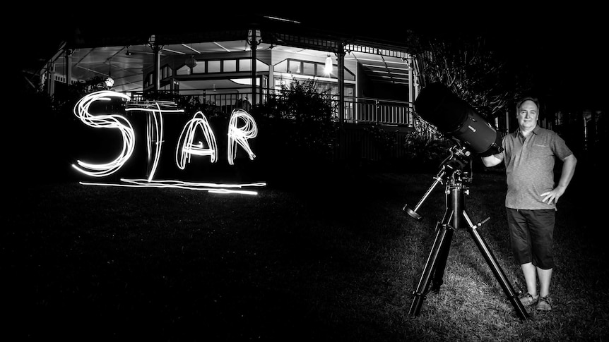 In otherwise darkness, a man leans against an amateur astronomer's telescope, with the word 'STAR' spelled out next to him.
