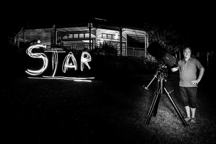 In otherwise darkness, a man leans against an amateur astronomer's telescope, with the word 'STAR' spelled out next to him.