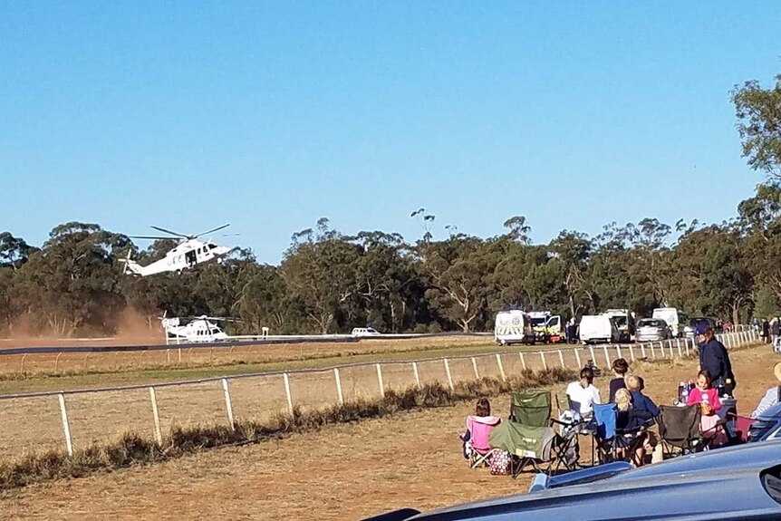 A helicopter landing at a racecourse while a small group of people watch on.