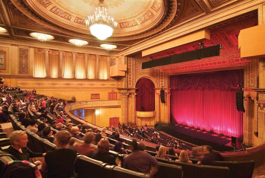 The inside of an opulent theatre where people and sitting down and looking at a red velvet curtain.
