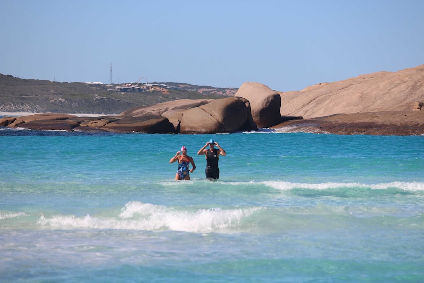 Two swimmers stand in the water near rocks on the coast