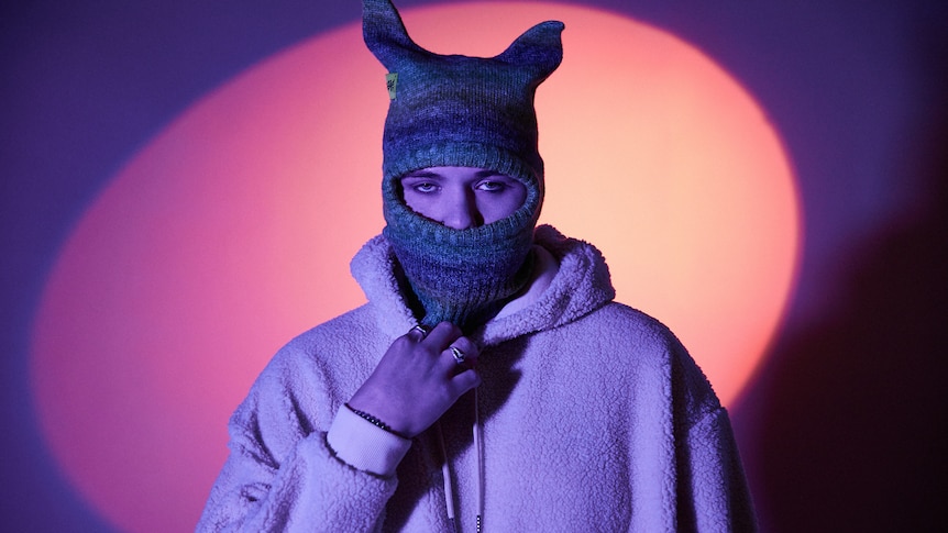 YNG Martyr in a fluffy white jumper and knitted balaclava with rabbit ears against a spotlight backdrop