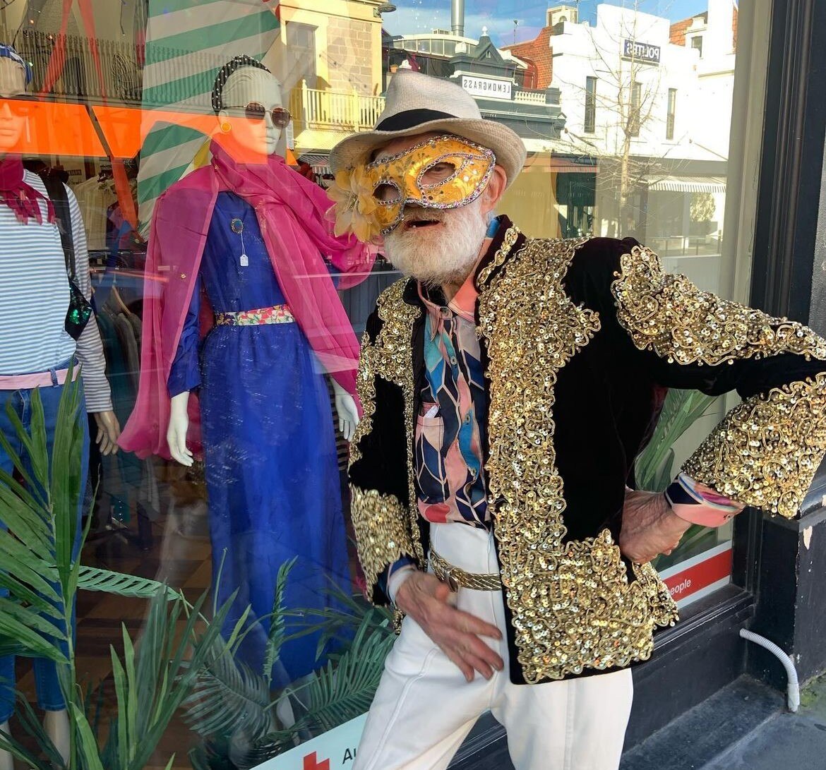 man in gold masquerade mask and sequined black and gold jacket with white hat posing outside a fashion store