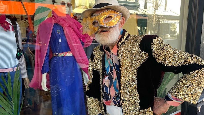 man in gold masquerade mask and sequined black and gold jacket with white hat posing outside a fashion store