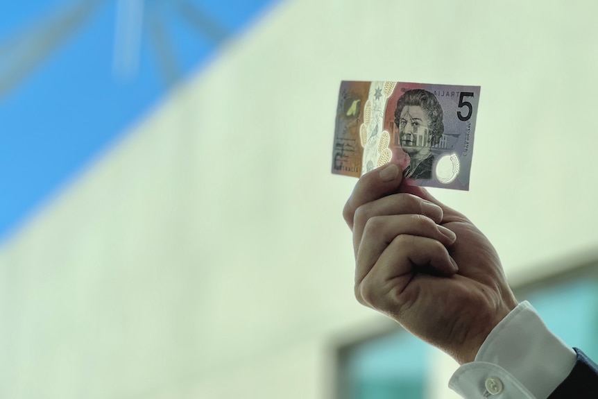A hand holds a $5 note, the spire of Parliament House just visible in the blurred background.