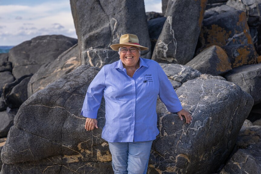 Wide shot of woman in denim shirt and hat leaning on large granite rocks beside the beach, smiling 