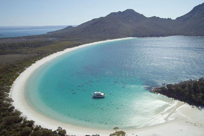 View of a curved bay with bright blue water, white sand and green trees behind the beach