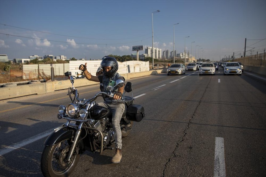 A man wearing jeans and t-shirt rides a motorbike down an Israel street gesturing a thumbs up