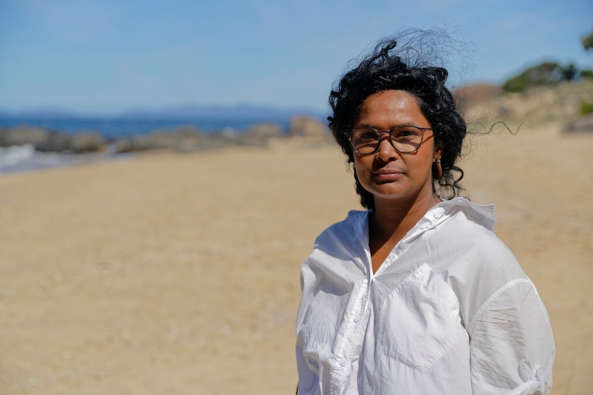 A woman with glasses wearing a white linen shirt standing on a beach.