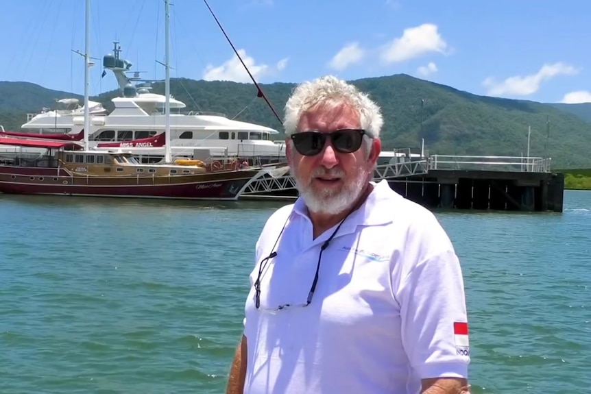 An older, bearded man with snowy hair stands in front of a some large leisure ships docked at a wharf on a sunny day.