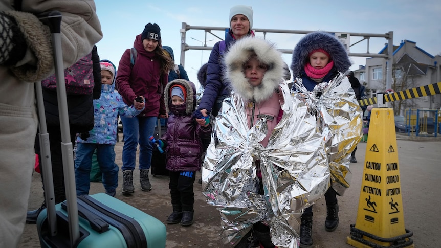 Children wearing foil blankets walk with their mothers on a road.