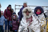 Children wearing foil blankets walk with their mothers on a road.