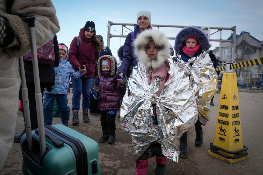 Children wearing foil blankets walk with their mothers on a road.