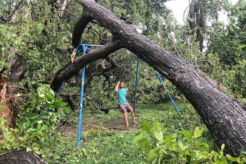 A man swings on a swingset pinned by a fallen tree in Darwin after Cyclone Marcus.