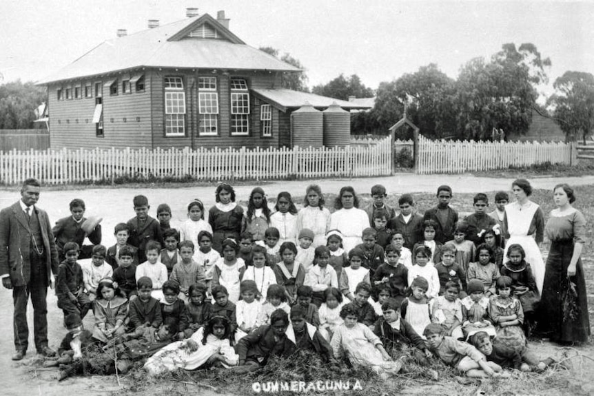 Black and white photo of a teacher and large group of students outside a weatherboard schoolhouse.