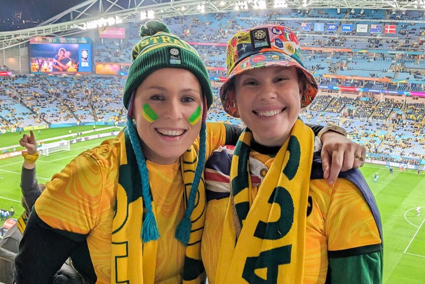 Two women decked out in Matildas merch grin in a stadium.