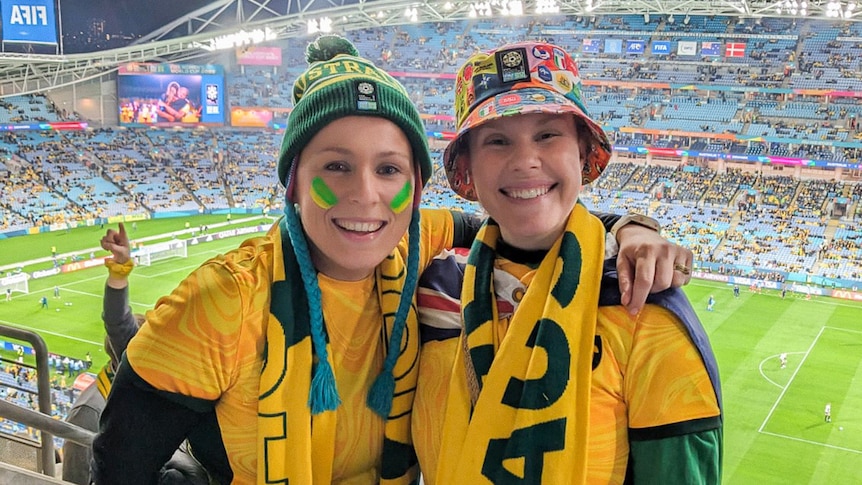 Two women decked out in Matildas merch grin in a stadium.