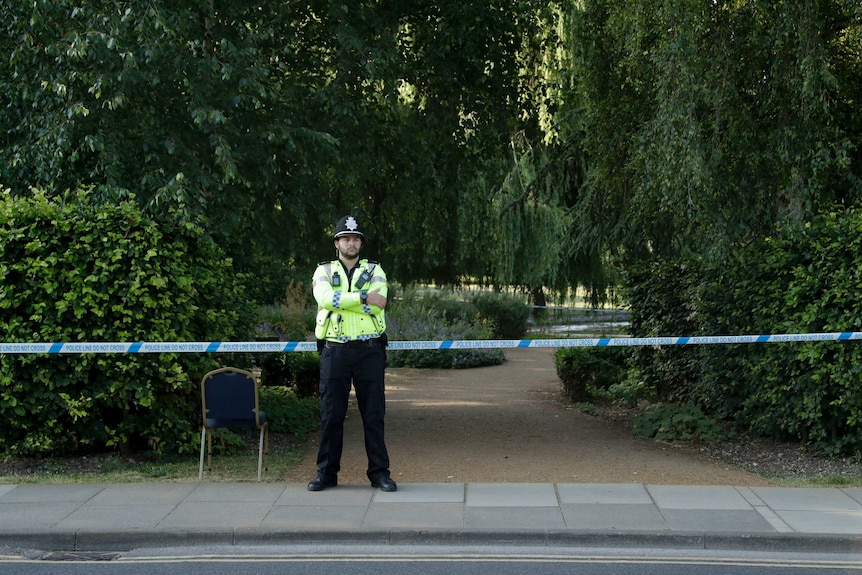 A police officers stands in front of police tape, which is closing off a park.