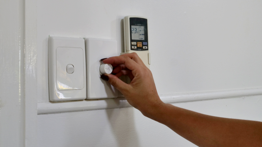 A hand turning a fan on, light switch and air con remote in the shot 