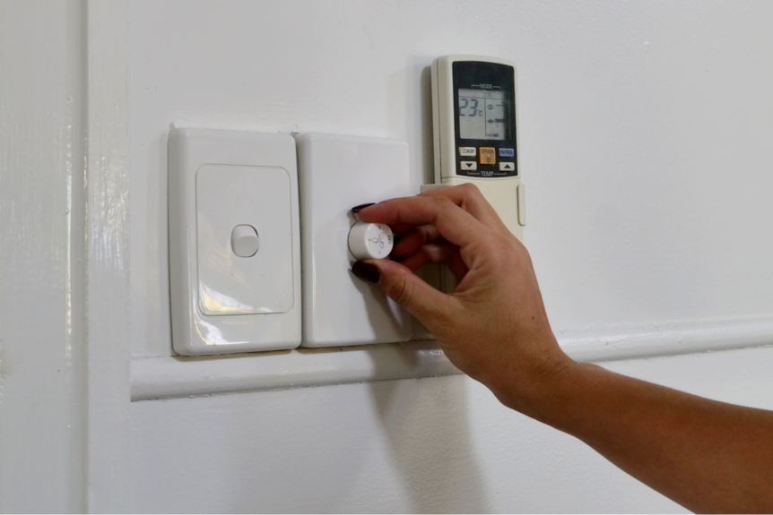 Una mano enciende un ventilador, un interruptor de luz y un control remoto del aire acondicionado en la toma 