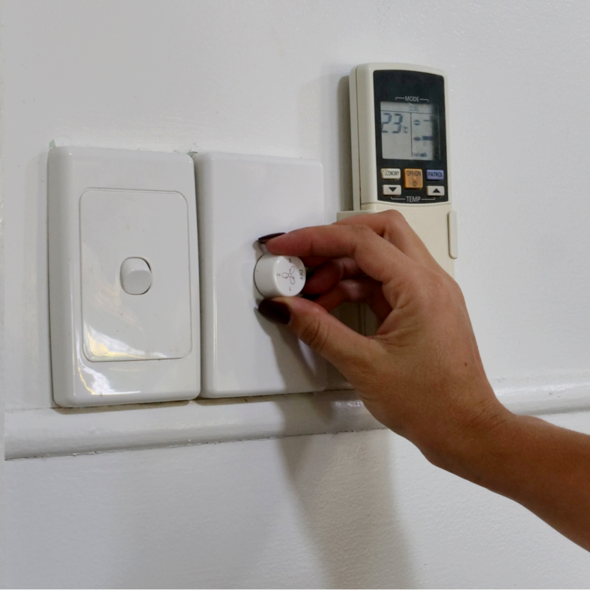 A hand turning a fan on, light switch and air con remote in the shot 