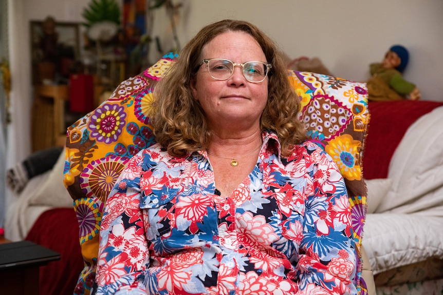 A woman wearing a bright flowery blouse gets sits on a chair. 
