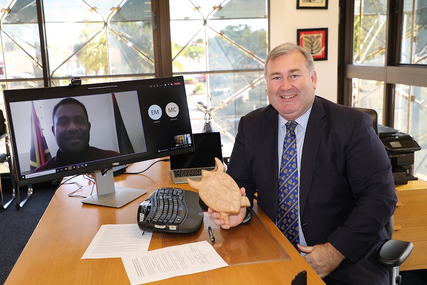 A middle aged white man smiles for the camera sitting next to a computer screen where he skype with a man from Vanuatu   