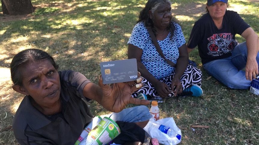 A woman sitting on grass holds a cashless welfare card up to the camera with two people nearby.