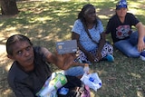 A woman sitting on grass holds a cashless welfare card up to the camera with two people nearby.