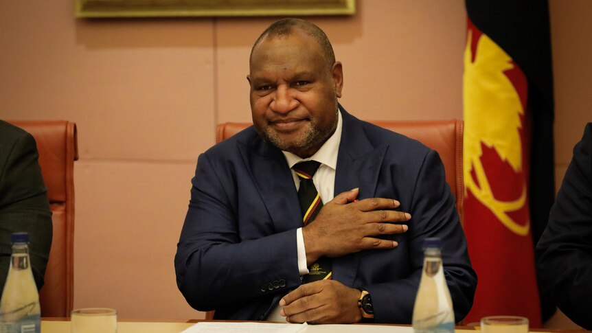 James Marape, wearing a suit and tie with red-yellow stripe down it, puts one hand to his chest