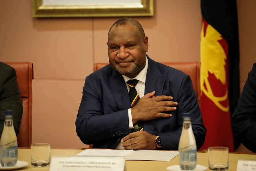 James Marape, wearing a suit and tie with red-yellow stripe down it, puts one hand to his chest