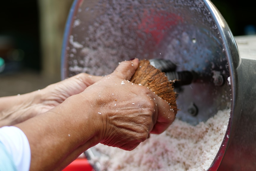 woman's hands holding a coconut on a motorised scraper with silver bowl in back.