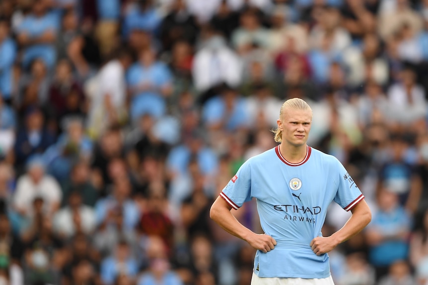 A male soccer player wearing light blue has his hands on his hips with a crowd behind him