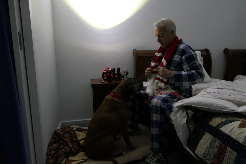A man sits on the edge of his bed in his PJs with a dog looking at him.