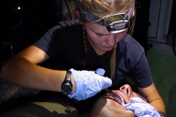 A teen girl wearing a headlamp and latex gloves injects the bums of a man wearing a military shirt.