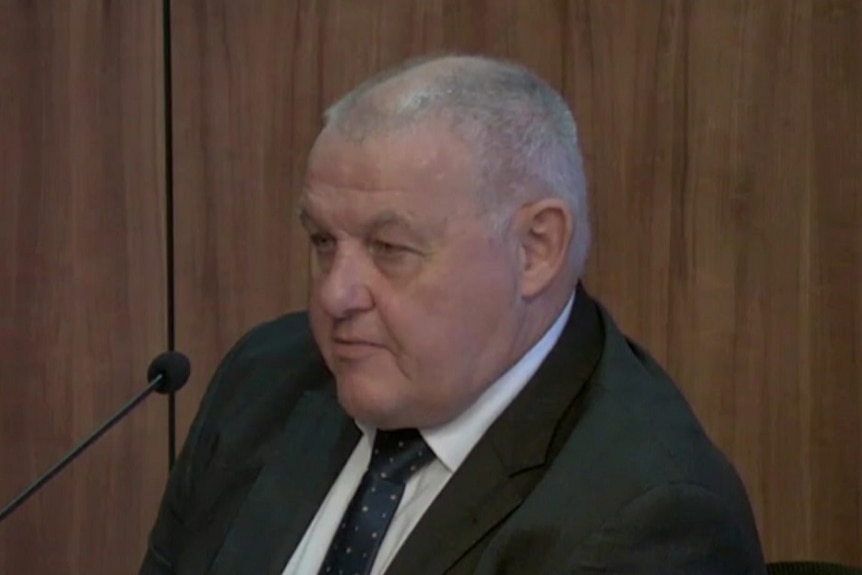 Ron Iddles, an older man with short grey hair, wearing a black suit and tie sitting down looking off to the left in a courtroom