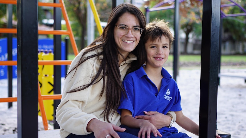 A young mother with long brown hair and glasses sitting on a playground with her young son