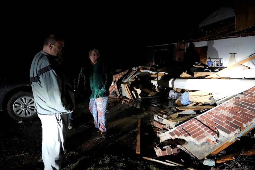 Two people standing next to a flattened house and its debris