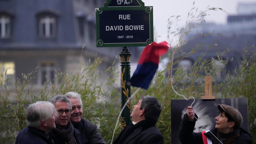 A sign says Rue David Bowie and a woman holds up a French flag 