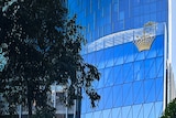 A golden logo of a crown made up of dots sits on the side of a blue-tinted glass building.