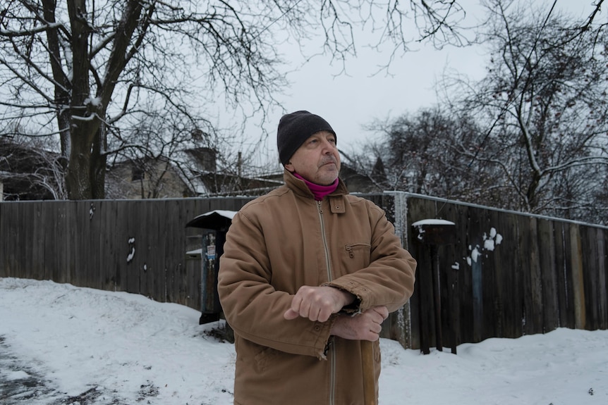 An older man in a brown coat stands in front of freshly shovelled snow on a street in Ukraine.