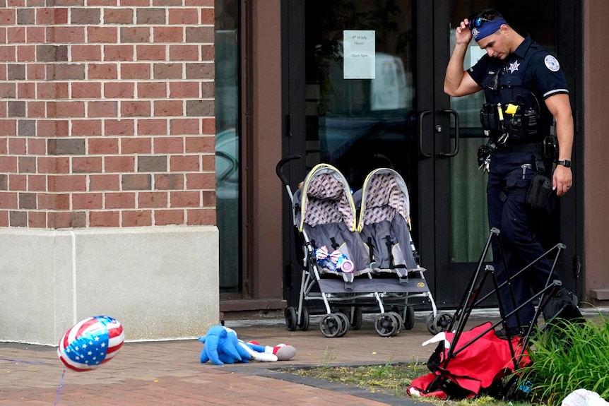 A police officer looks down, American flag balloons on the ground, a stroller in the background