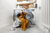 A woman sits on the ground in front of a washing machine and dryer, sorting her clothes.