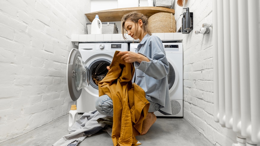 A woman sits on the ground in front of a washing machine and dryer, sorting her clothes.