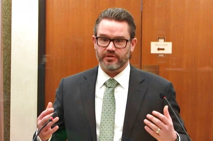 You view a man in a dark suit with a green tie speaking into two small microphones while gesticulating with his hands.