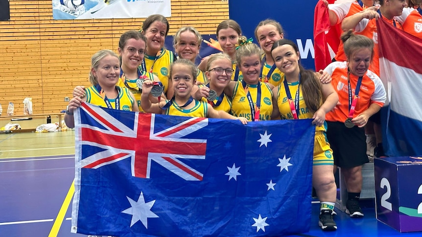 A group of short statured women wearing green and gold basketball singlets and holding an Australian flag smile at camera.