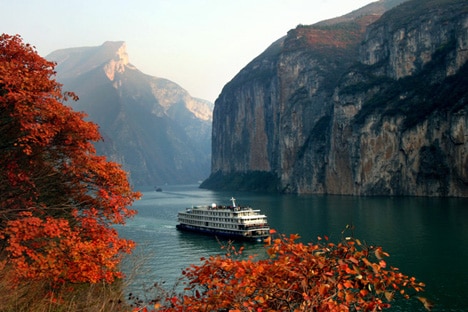 A large cruise ship passes along a large river under towering sheer cliffs topped with forest.