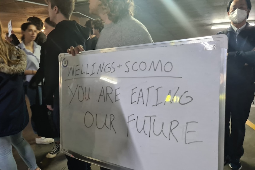 A woman holds a small whiteboard with the words Wellings + Scomo, You are eating our future