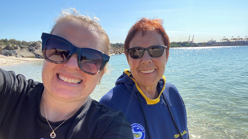 Natalie with her mother standing in front of a beach with sunglasses on.