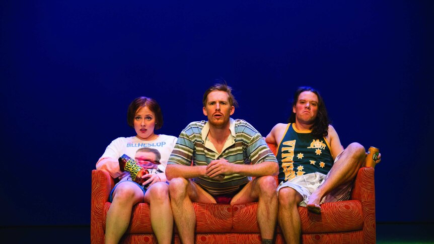 Three people sit on a couch watching tv with junk food on stage in Muriel's Wedding the musical